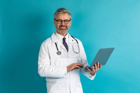 Telemedicine, telecommunication in healthcare. Happy cheerful attractive mature european man therapist in workwear using modern pc laptop on blue studio background, copy space