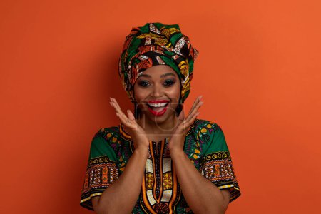 Téléchargez les photos : Portrait d'une belle jeune femme noire excitée et étonnée portant un costume traditionnel et un maquillage lumineux sur fond de studio orange, s'exclamant et se tenant la main par le visage - en image libre de droit