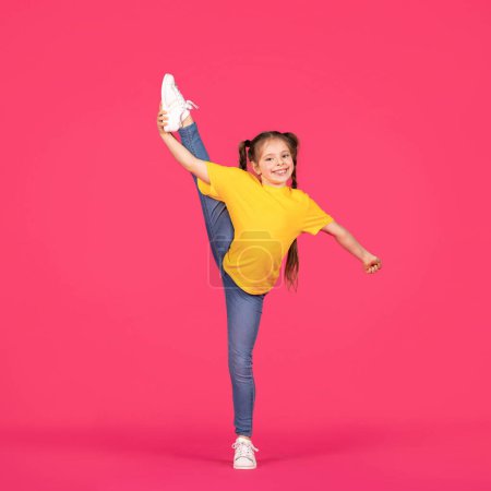 Photo for Cute little gymnast girl raising her leg in twine while posing over pink studio background, cheerful preteen female child in casual clothes making gymnastics, doing stretching exercises, full length - Royalty Free Image
