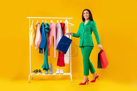Photo for Shopping and seasonal sales. Happy stylish woman walking with shopper bags near clothing rail on yellow studio background, full length shot, free space - Royalty Free Image