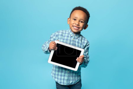 Téléchargez les photos : Apps et enseignement à distance pour les enfants. Heureux garçon afro-américain montrant tablette avec écran blanc et souriant à la caméra, posant isolé sur fond bleu, maquette - en image libre de droit