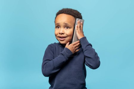 Téléchargez les photos : Portrait d'un adorable garçon afro-américain parlant sur un téléphone portable avec un ami, isolé sur fond bleu, plan studio, espace de copie gratuit - en image libre de droit