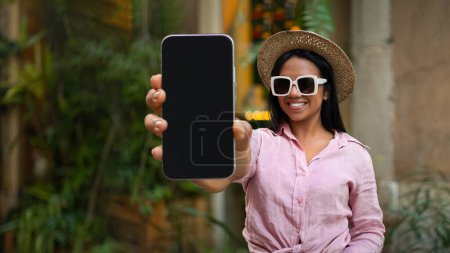 Photo for Happy millennial african american female tourist in hat and sunglasses shows phone with empty screen, enjoy travel in city, recommends app for trip blog outdoor. Vacation and journey at free time - Royalty Free Image