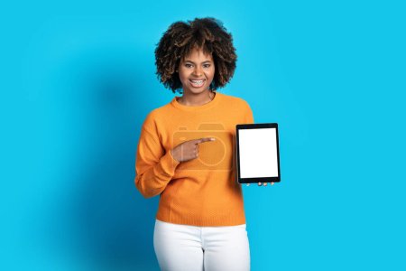 Foto de Almohadilla digital de gadget moderno con pantalla blanca vacía en mano de mujer negra, alegre y atractiva dama afroamericana apuntando a la tableta con espacio vacío de maqueta para publicidad, fondo azul - Imagen libre de derechos