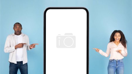 Photo for Happy african american spouses pointing at huge cellphone with blank screen, advertising new application or website, standing over blue background, mockup, banner - Royalty Free Image