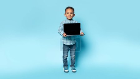 Photo for Adorable black boy showing laptop with blank screen, standing on blue studio background, mockup for design, full length. Happy male child holding computer with template for website - Royalty Free Image