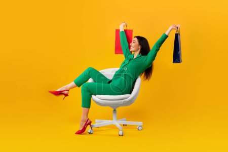 Photo for Happy contented shopaholic woman sitting on chair and raising hands with shopping bags isolated over yellow studio background, full length, free space - Royalty Free Image