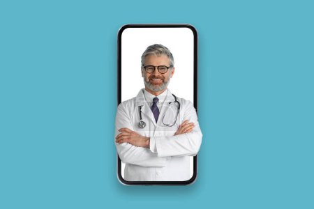 Friendly cheerful handsome grey-haired middle aged european man doctor with arms crossed on chest in big cell phone screen, isolated on blue background. Telemedicine, telecare concept