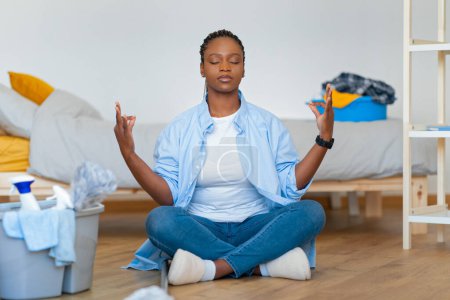 Photo for Household chores, home cleaning. Peaceful young black woman in homewear housewife sitting in lotus pose with closed eyes on floor among cleaning tools, meditating, untidy bedroom interior - Royalty Free Image