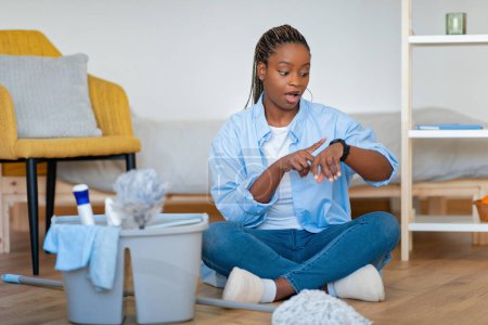 Photo for Stressed overwhelmed busy young african american lady in homewear sitting on floor in bedroom with basket full of cleaning tools, checking smart watch, have no time for chores - Royalty Free Image