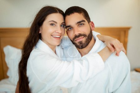 Téléchargez les photos : Couple aimant étreignant portant des peignoirs blancs posant assis dans la chambre à coucher à l'intérieur, souriant à la caméra. Portrait de heureux conjoints mixtes. Amour et relation romantique Concept - en image libre de droit