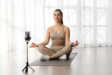 Photo for Peaceful attractive young woman yoga instructor sitting in lotus position, meditating and recording video for virtual yoga class, using cell phone set on tripod, fitness studio interior - Royalty Free Image