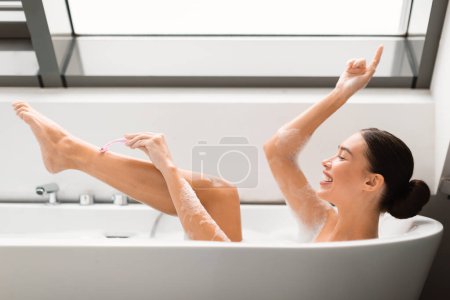 Téléchargez les photos : Happy Woman Rasage des jambes en utilisant le rasoir de sécurité dans la baignoire, profiter de la routine de dépilation prenant bain dans la salle de bain moderne à la maison. Épilation et soins du corps Cosmétiques Concept. Vue latérale - en image libre de droit