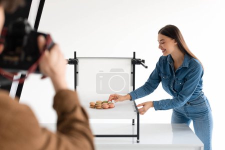 Photo for Professional male photographer with photocamera taking photos of sweet food macarons on white platform while his female assistant standing nearby and helping - Royalty Free Image