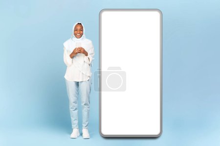 Photo for Cool app. Happy black muslim woman posing near huge smartphone with blank screen and smiling at camera, using mobile phone, standing on blue studio background, mock up, full body length - Royalty Free Image