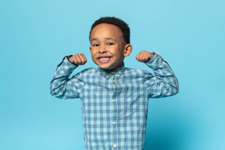 Photo for Funny black boy showing biceps and smiling at camera, small african american male child demonstrating his power and strength while posing over blue background - Royalty Free Image