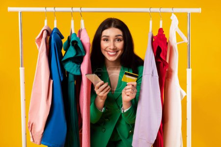 Photo for Excited young female customer holding cellphone and credit card, posing near clothing rail over yellow studio background, smiling at camera. Happy shopaholic buying clothes online - Royalty Free Image