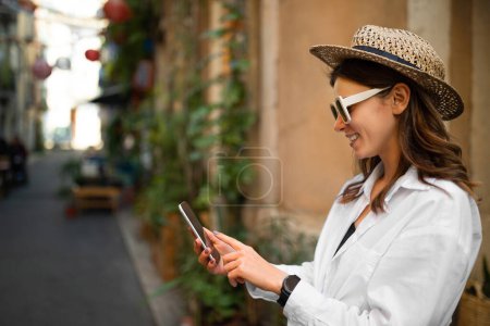 Photo for Smiling young european woman tourist in hat, sunglasses, typing on smartphone, looking at online map in city, outdoor. Tourism, app for travel blog, vacation journey, social media and active lifestyle - Royalty Free Image