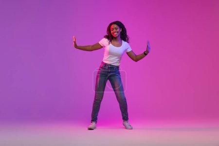 Téléchargez les photos : Portrait De Joyeuse Femme Afro-Américaine Danse Au Néon Lumière Sur Dégradé Violet Arrière-plan En Studio, Pleine Longueur Prise De Vue De Joyeux Jeune Femme Noire S'amusant Et Souriant À La Caméra, Espace De Copie - en image libre de droit