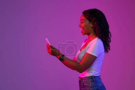 Photo for Mobile Offer. Excited Black Woman In Neon Light Looking At Smartphone Screen, Cheerful Young African American Female Holding Mobile Phone, Playing Online Games Or Messaging, Side View With Copy Space - Royalty Free Image