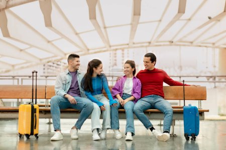 Photo for Vacation With Friends. Two Happy Couples Tourists With Suitcases Sitting Waiting For Flight In Modern Airport Terminal Indoors. Group Of Young People Traveling Together. Tourism And Transportation - Royalty Free Image