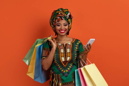 Téléchargez les photos : Positif joyeux jolie jeune femme noire dans les achats nationaux de costumes africains en ligne, tenant achats sacs en papier colorés, en utilisant un smartphone, isolé sur fond de studio orange - en image libre de droit