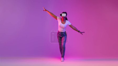 Foto de Mujer negra alegre en auriculares VR explorando actividades del ciberespacio, caminando en luz de neón sobre fondo de estudio púrpura, Emocionado equilibrio femenino afroamericano al hacer pasos, Copiar el espacio - Imagen libre de derechos