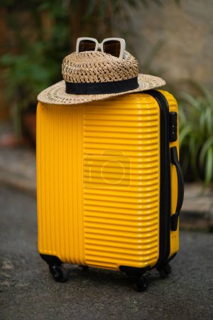 Photo for Yellow suitcase with hat and sunglasses on street, accessories for trip for tourist vertical, close up, nobody, outdoor. Travel vacation prepare, summer holiday and active lifestyle - Royalty Free Image