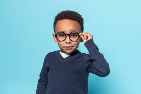 Téléchargez les photos : Petit garçon afro-américain intelligent en uniforme scolaire touchant des lunettes et regardant la caméra, debout sur fond de studio bleu, espace libre - en image libre de droit
