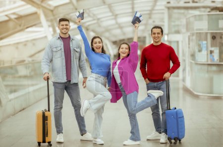Téléchargez les photos : Offre de billets pas chers. Deux couples d'amis partent en vacances tenant des laissez-passer d'embarquement, debout avec des valises de voyage souriant à la caméra posant ensemble à l'aéroport à l'intérieur. Longueur totale - en image libre de droit
