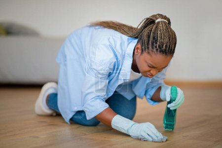 Photo for Annoyed young black woman housewife or maid standing on knees, erasing removing dirt stain from wooden floor, using cleaning spray and duster cloth, home imterior, copy space - Royalty Free Image