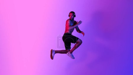 Der athletische Afroamerikaner, der in der Luft posiert und Musik hört, trägt drahtlose Kopfhörer über lila Neon-Studiohintergrund. Workout Playlist Konzept. Panorama