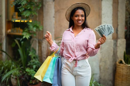 Photo for Cheerful african american woman in casual and hat with many bags of purchases hold dollars, enjoy shopping in city, outdoor. Huge sale, money for buy and fashion at vacation, buying trip - Royalty Free Image