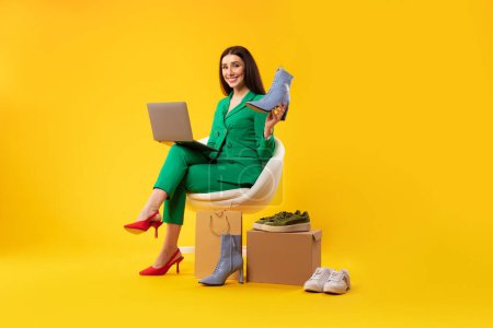 Téléchargez les photos : Jeune styliste féminine avec ordinateur portable et de nouvelles chaussures, assise sur une chaise sur fond de studio jaune, pleine longueur, espace libre. Achats en ligne et concept de consultation de style - en image libre de droit