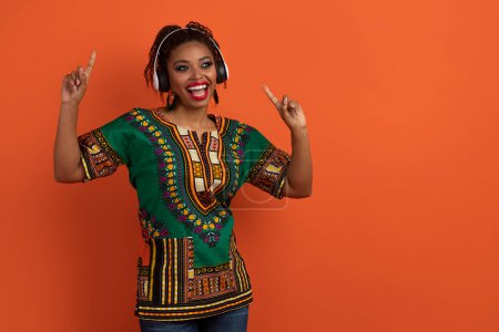 Téléchargez les photos : Drôle heureuse jeune femme afro-américaine en costume national traditionnel s'amuser sur fond de studio orange, en utilisant des écouteurs sans fil, écouter de la musique, danser, chanter, copier l'espace - en image libre de droit