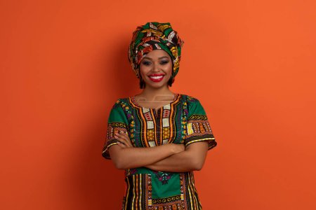 Foto de Alegre hermosa mujer afroamericana joven en traje tradicional colorido posando con los brazos cruzados en el pecho sobre fondo de estudio naranja, sonriendo a la cámara. Mujeres, mujeres, feminidad - Imagen libre de derechos