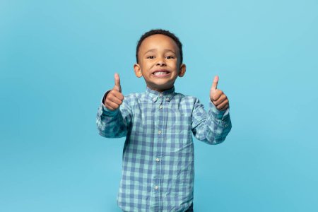 Photo for Portrait of cute black boy showing thumbs up and smiling at camera while posing over blue background, studio shot. Happy african american male child gesturing sign of approval - Royalty Free Image