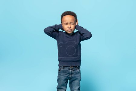 Photo for Unhappy black little kid boy closing eyes and ears being annoyed with noise wanting silence and calm atmosphere, standing isolated over blue studio background - Royalty Free Image