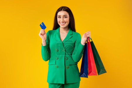 Téléchargez les photos : Portrait d'une femme élégante heureuse tenant des sacs à provisions et une carte de crédit, posant après avoir magasiné en magasin sur fond jaune, prise de vue en studio - en image libre de droit