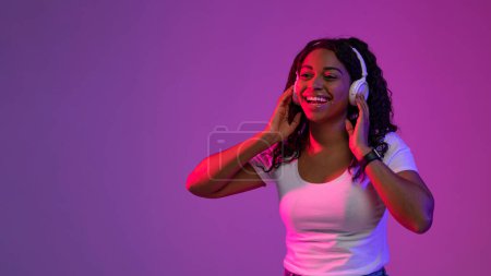 Téléchargez les photos : Nouvelle playlist. Joyeux jeune femme noire portant des écouteurs sans fil debout dans la lumière au néon sur fond violet Studio, Happy African American Femme écoutant de la musique préférée, Copier l'espace - en image libre de droit