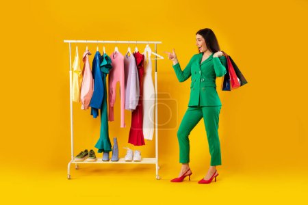 Photo for Shopaholic. Excited stylish woman pointing finger at clothing rail, shopping and choosing new clothes, standing on yellow studio background, full length - Royalty Free Image