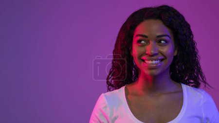 Téléchargez les photos : Portrait de jeune femme noire pensive regardant à côté avec intérêt, femme afro-américaine rêveuse considérant des options tout en se tenant debout dans la lumière au néon sur fond violet, Panorama, Copier l'espace - en image libre de droit