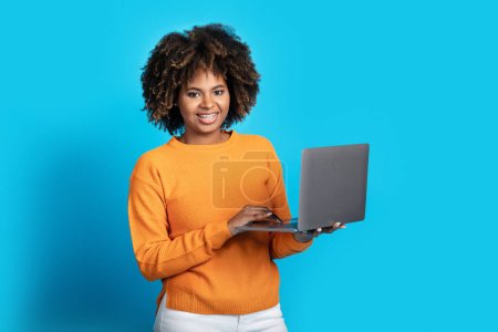 Nice online offer, app. Positive attractive young black lady with bushy hair shopping online, holding modern laptop, smiling at camera, posing on blue studio background, copy space