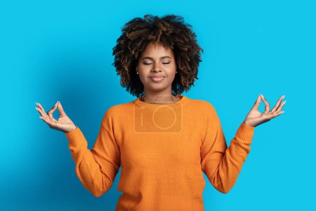 Téléchargez les photos : Paisible jeune femme noire détendue aux cheveux touffus méditant les yeux fermés sur fond de studio bleu, tenant la main en geste zen. Relaxation, soulagement du stress, concept de pleine conscience - en image libre de droit