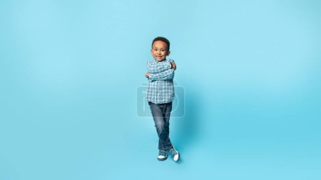 Foto de Foto completa de niño afroamericano posando con los brazos cruzados, mirando y sonriendo a la cámara sobre fondo azul, panorama con espacio libre. concepto de belleza y estilo para niños - Imagen libre de derechos