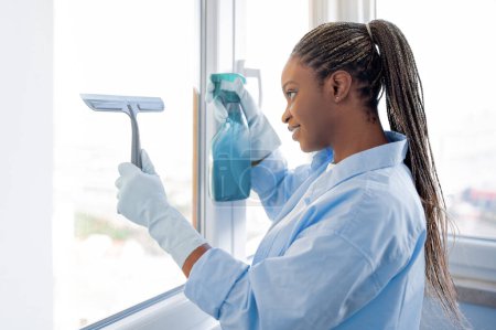 Photo for Professional housekeeping, cleaning service. Cheerful attractive young black woman housekeeper cleaning windows at apartment, using spray and cleaner, wearing rubber gloves, side view, copy space - Royalty Free Image