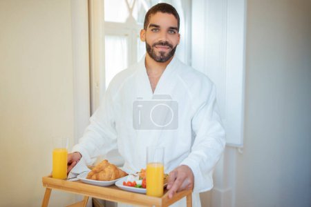 Téléchargez les photos : Petit déjeuner romantique au lit. Beau Moyen-Orient homme tenant plateau de table avec croissants, salade et jus d'orange souriant à la caméra debout dans la chambre d'hôtel à l'intérieur, portant un peignoir blanc - en image libre de droit