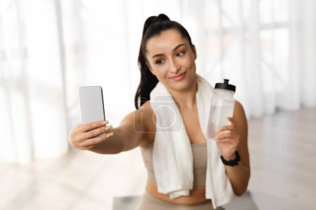 Photo for Friendly cheerful athletic brunette young woman in sportswear fitness blogger taking selfie on cell phone, female yoga instructor showing bottle of water, recommend water drinking during workout - Royalty Free Image