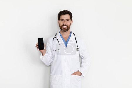 Foto de Sonriente médico masculino en uniforme que demuestra el teléfono inteligente en blanco en su mano mientras está parado cerca de la pared blanca en la clínica, Hombre guapo terapeuta de capa blanca que recomienda la aplicación médica móvil, Mockup - Imagen libre de derechos