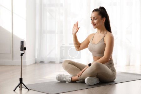Téléchargez les photos : Cours de yoga virtuel concept. Professeur de yoga millénial belle femme en tenue de sport serrée assise sur un tapis de fitness, agitant la caméra du smartphone, saluant les étudiants, copiant l'espace - en image libre de droit